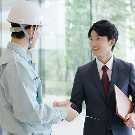 作業員とスーツ姿のビジネスマンが資料を確認しながら会話している様子