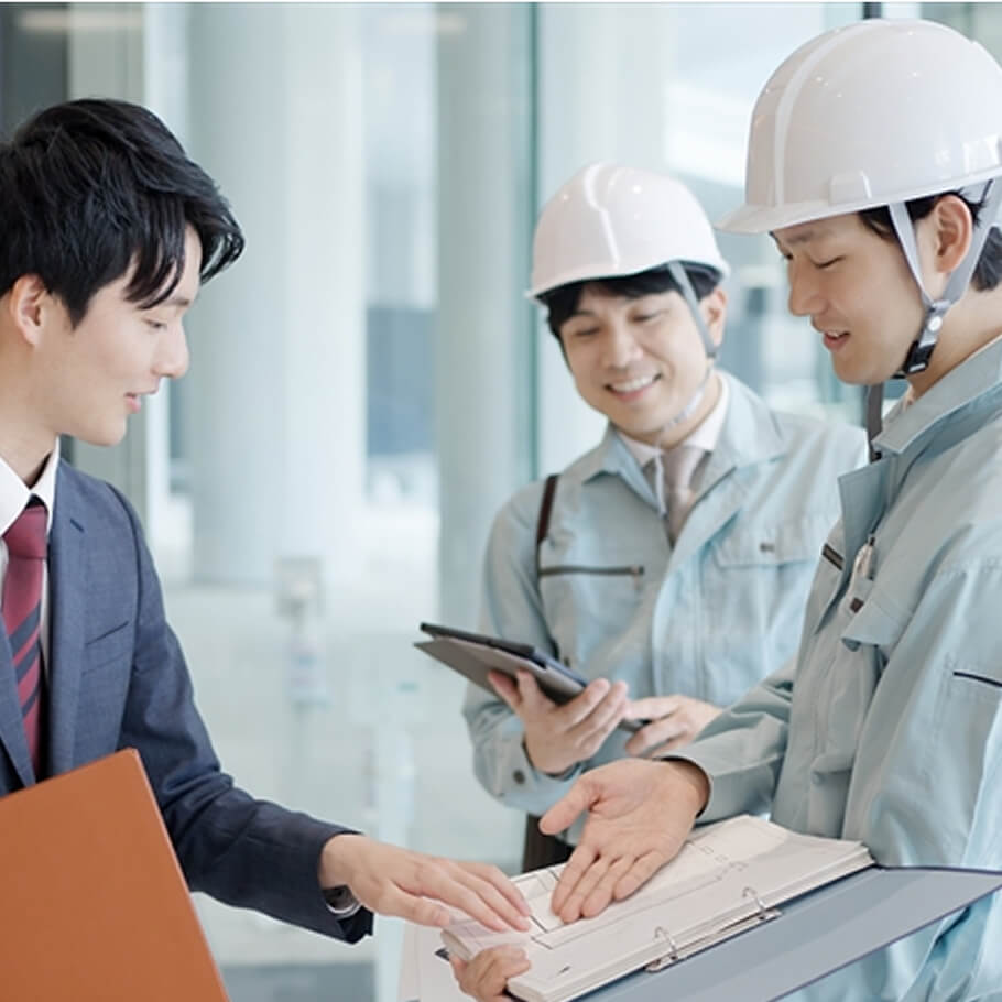 スーツ姿の担当者と作業員が現場で報告書を見ながら打ち合わせをしている様子